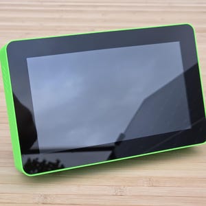 May include: A rectangular tablet with a black screen and a bright green border. The screen reflects the sky and surrounding environment. The device is placed on a light brown wooden surface.