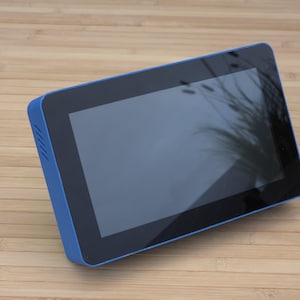 Könnte beinhalten: Ein Tablet mit blauem Rahmen und schwarzem Bildschirm liegt auf einer hellbraunen Holzoberfläche. Der Bildschirm spiegelt ein verschwommenes Bild wider. Der Rahmen des Tablets hat eine matte Oberfläche, und der Bildschirm ist bündig mit dem Rahmen.