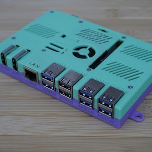 Puede incluir: Un dispositivo electrónico turquesa y morado con múltiples puertos USB 3.0, un puerto LAN y ranuras de ventilación. El dispositivo tiene un ventilador y tiene forma rectangular. Los puertos son plateados.