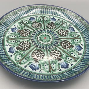 May include: A large, round, white ceramic plate with a blue and green floral design. The design features stylized pinecones and leaves.