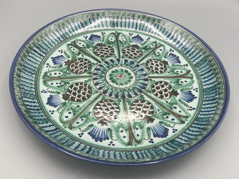 May include: A large, round, white ceramic plate with a blue and green floral design. The design features stylized pinecones and leaves.