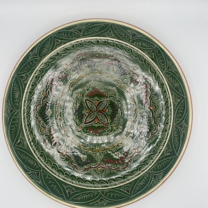 Può includere: Un piatto rotondo in ceramica verde con un intricato disegno dipinto a mano. Il piatto presenta un motivo floreale centrale rosso e bianco, circondato da motivi concentrici e un bordo dettagliato. Il bordo del piatto è beige chiaro.