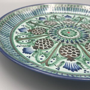 May include: A large, round ceramic plate with a blue and green floral design. The plate features a central motif of a stylized flower surrounded by eight pinecone-like shapes.