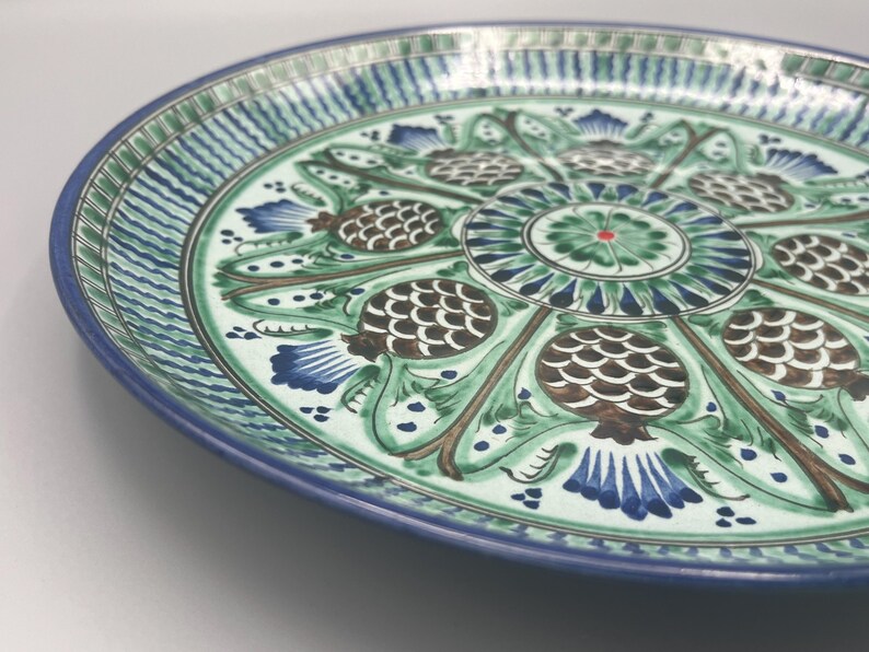 May include: A large, round ceramic plate with a blue and green floral design. The plate features a central motif of a stylized flower surrounded by eight pinecone-like shapes.