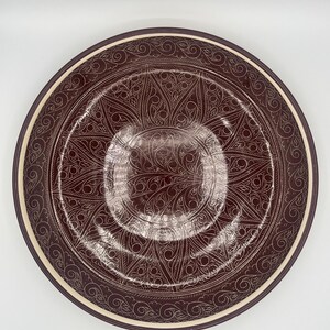 May include: A round, decorative plate with a dark brown center and a cream-colored rim. The plate features an intricate, white, floral pattern. The rim is a darker shade of brown. The plate is likely ceramic or porcelain.