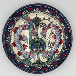 Può includere: Un piatto rotondo in ceramica con un bordo blu e bianco e un disegno centrale di un'anfora stilizzata verde e blu con bacche e foglie rosse.