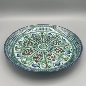 May include: A large, round, ceramic plate with a green and blue floral design. The plate has a white background and features a central motif of a stylized flower with eight petals. The petals are decorated with brown and green details. The plate is surrounded by a border of green and blue stripes.