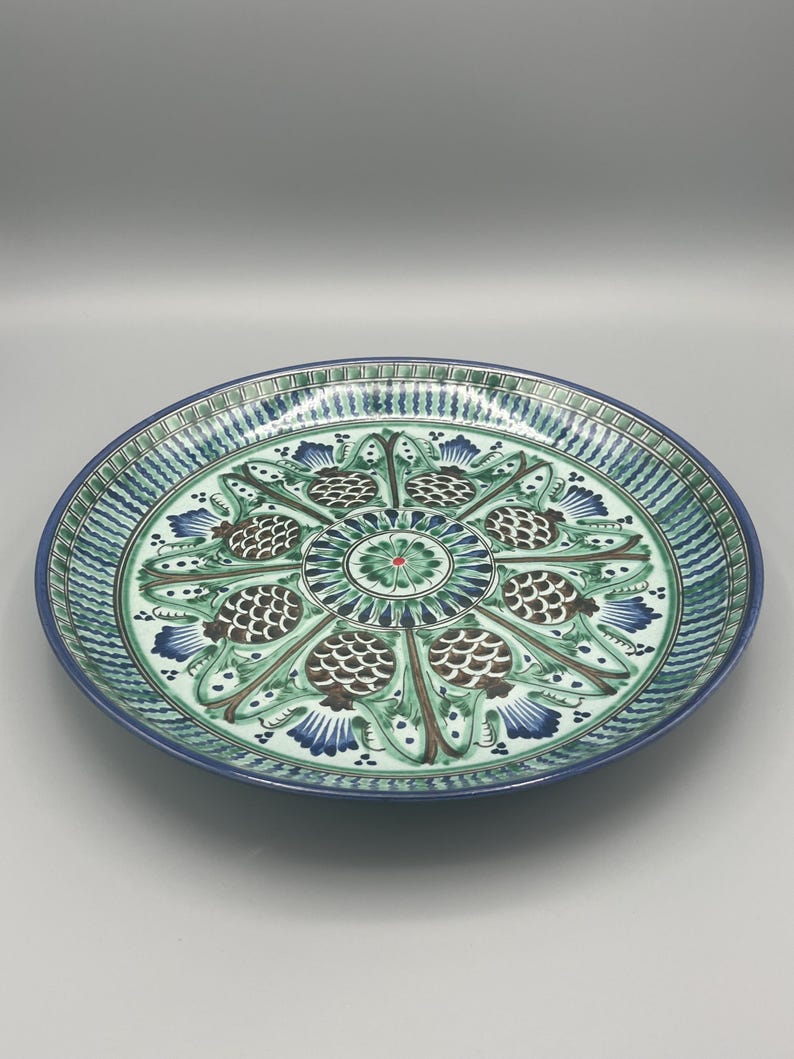 May include: A large, round, ceramic plate with a green and blue floral design. The plate has a white background and features a central motif of a stylized flower with eight petals. The petals are decorated with brown and green details. The plate is surrounded by a border of green and blue stripes.