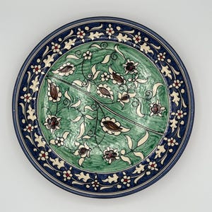 Può includere: Un piatto rotondo in ceramica con sfondo verde e un design floreale blu e bianco. Il piatto ha un bordo decorativo con fiori rossi e bianchi.