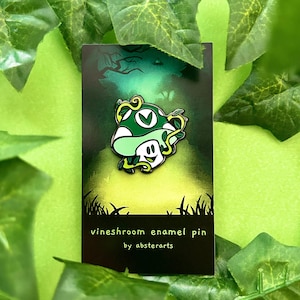 Puede incluir: Pin de esmalte verde de un hongo con enredaderas que crecen a su alrededor. El hongo tiene una cara blanca con una sonrisa y un corazón verde en su sombrero. El texto "vineshroom enamel pin by absterarts" está impreso en la tarjeta.