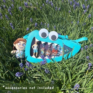 May include: A bright turquoise monster-shaped crossbody bag with large white googly eyes and a wide mouth featuring white triangular teeth. The bag has a clear front panel displaying various small character figures and a strap attached to the side.
