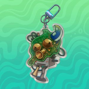 Puede incluir: Un amuleto verde y dorado con purpurina con un personaje de dibujos animados que sostiene una trompeta. El amuleto está unido a un llavero plateado.