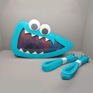 May include: A bright turquoise monster-shaped crossbody bag featuring two large white googly eyes, a wide mouth with white triangular teeth, and a transparent mesh front panel. The bag includes two matching turquoise adjustable shoulder straps.