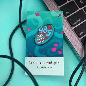 Könnte beinhalten: Eine blaue und pinke Emaille-Anstecknadel mit einem Cartoon-Keim mit der Nummer 985 darauf. Die Anstecknadel befindet sich auf einer Karte mit dem Text "jern enamel pin by absterarts".