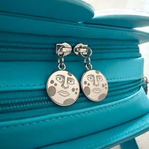 May include: A bright turquoise bag featuring two silver-toned circular zipper pulls. Each pull is engraved with a detailed illustration of a human-like face with large eyes and circular markings on the cheeks, suspended by a small metal chain link.