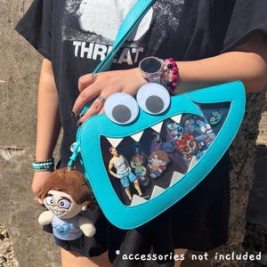 May include: A bright turquoise crossbody bag shaped like a monster mouth with large googly eyes and white triangular teeth. The bag features a clear front panel displaying various character charms and a plush doll keychain hanging from the side.