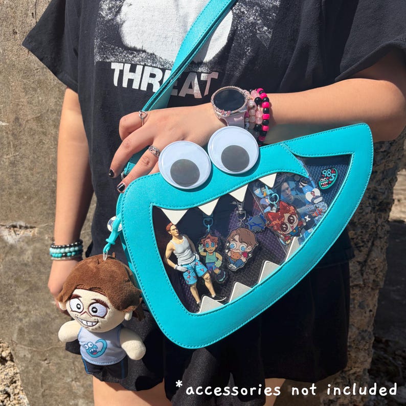 May include: A bright turquoise crossbody bag shaped like a monster mouth with large googly eyes and white triangular teeth. The bag features a clear front panel displaying various character charms and a plush doll keychain hanging from the side.