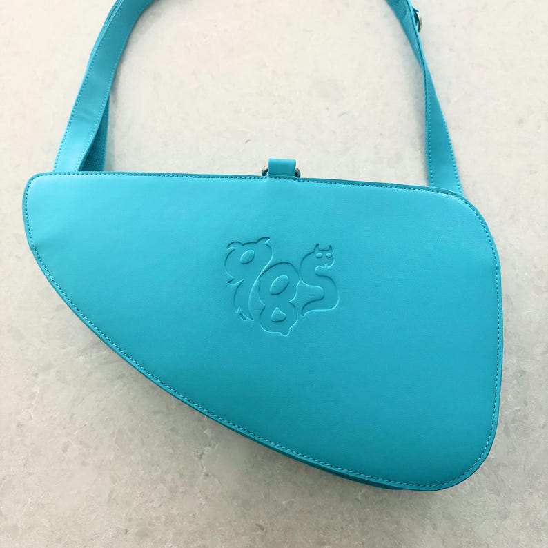 May include: A bright turquoise shoulder bag with an asymmetrical, curved silhouette and a long adjustable strap. The front features a debossed graphic logo design in the center. The bag has a smooth finish and a single top closure loop.