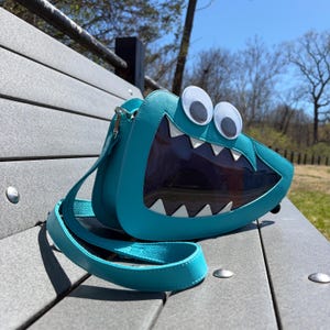 May include: A bright turquoise crossbody bag shaped like a monster face, featuring two large white googly eyes on top and a wide, dark-tinted transparent mouth section lined with sharp white triangular teeth and a matching turquoise shoulder strap.