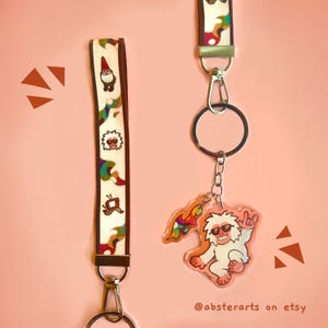 May include: Two lanyards and a keychain with a pink background. One lanyard is white with brown edges and colorful gnome and monster designs. The keychain features a clear, smiling yeti with sunglasses and a rainbow flame design.