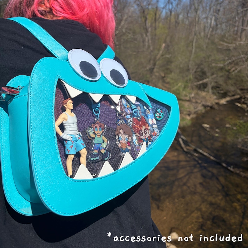 May include: A bright turquoise monster-shaped backpack with large white googly eyes, a wide mouth opening with white triangular teeth, and a dark purple mesh display window. The bag contains several small character figures and acrylic charms.