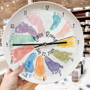 May include: A white ceramic clock featuring colourful footprints around the edge. The clock face displays the name "Calix Rome" and the date "July 22, 2024". Black clock hands show the time.