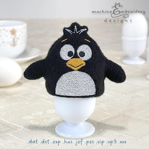 Puede incluir: Un calienta huevos de pingüino negro y blanco con pico y ojos amarillos. El calienta huevos está en un soporte de huevo blanco.