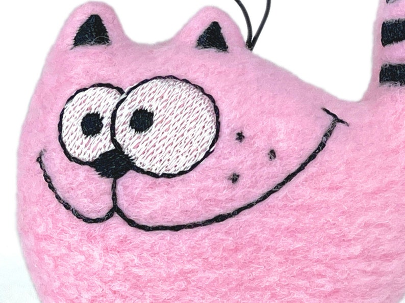 May include: A pink cat-shaped ornament with embroidered black details. The cat has large, expressive eyes, a smiling mouth, and black stripes on its tail. The ornament is made of a soft, fuzzy material.