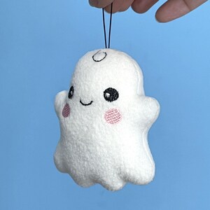 Puede incluir: Un peque&ntilde;o adorno de fantasma de peluche blanco con una textura suave y difusa. Presenta ojos negros bordados, una sonrisa negra sencilla y dos parches circulares rosados en las mejillas. Un cord&oacute;n negro fino est&aacute; unido a la parte superior.