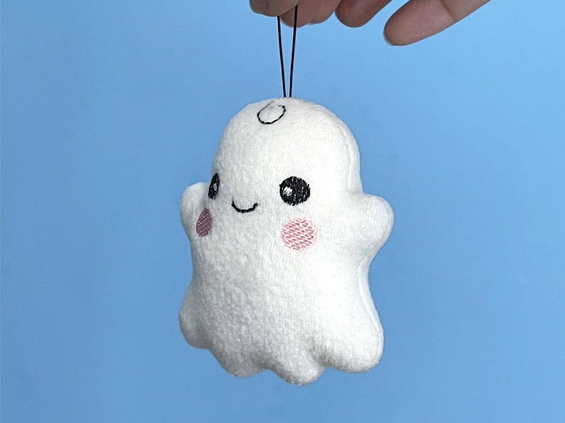 Puede incluir: Un peque&ntilde;o adorno de fantasma de peluche blanco con una textura suave y difusa. Presenta ojos negros bordados, una sonrisa negra sencilla y dos parches circulares rosados en las mejillas. Un cord&oacute;n negro fino est&aacute; unido a la parte superior.