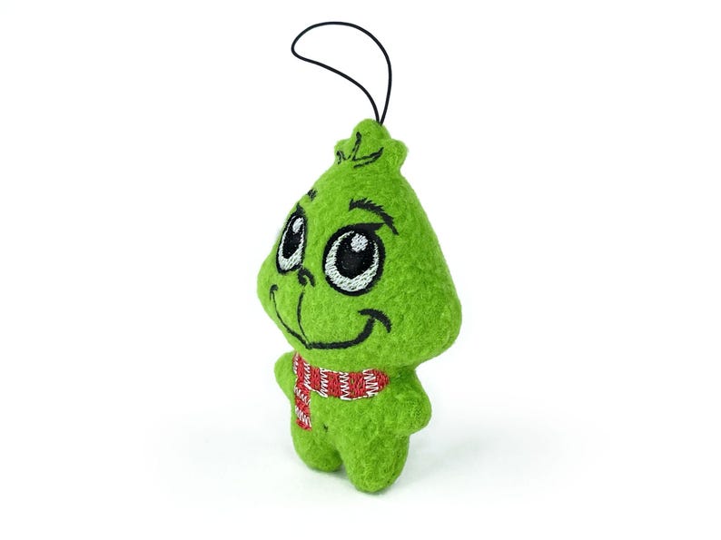 May include: Green plush ornament of the Grinch with a red and white striped scarf. The Grinch has a smiling face with large eyes and a black embroidered nose.