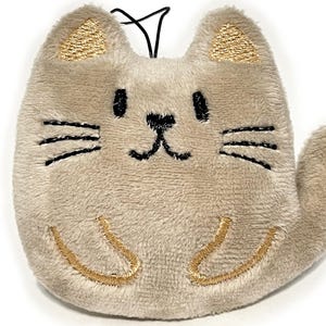 May include: A plush cat-shaped keychain in a light tan color. The cat has embroidered black whiskers, eyes, and a nose. The ears and paws are accented with gold embroidery. A black string is attached at the top.