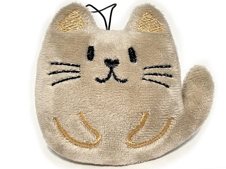 May include: A plush cat-shaped keychain in a light tan color. The cat has embroidered black whiskers, eyes, and a nose. The ears and paws are accented with gold embroidery. A black string is attached at the top.