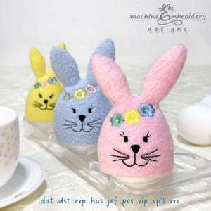 Könnte beinhalten: Drei Osterei-Wärmer in Form von Hasen in Blau, Gelb und Rosa. Jeder Hase trägt eine Blumenkrone und bestickte Gesichtszüge. Die Hasen sitzen auf einem durchsichtigen Plastiktablett. Der Text "machine embroidery designs" ist in der oberen rechten Ecke des Bildes sichtbar.