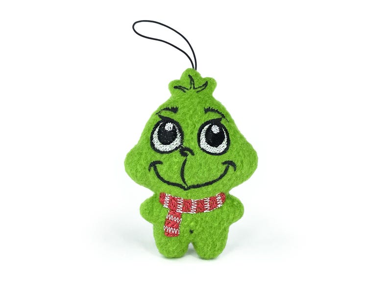 May include: Green plush ornament of the Grinch with a red and white striped scarf. The Grinch has a mischievous grin and large eyes.