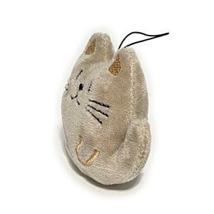 May include: A plush cat-shaped toy in a light beige color. The cat's face features embroidered black whiskers, eyes, and a smiling mouth. The ears are accented with gold thread. A black string is attached at the top.