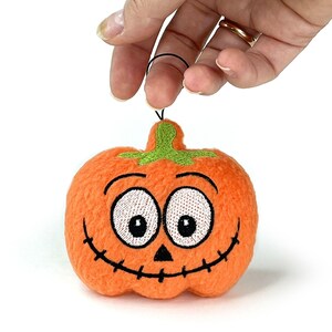 Puede incluir: Un adorno de calabaza naranja de felpa con textura difusa, que presenta ojos blancos bordados con pupilas negras, una peque&ntilde;a nariz triangular negra y una boca ancha cosida en negro. Un tallo verde bordado se encuentra en la parte superior.
