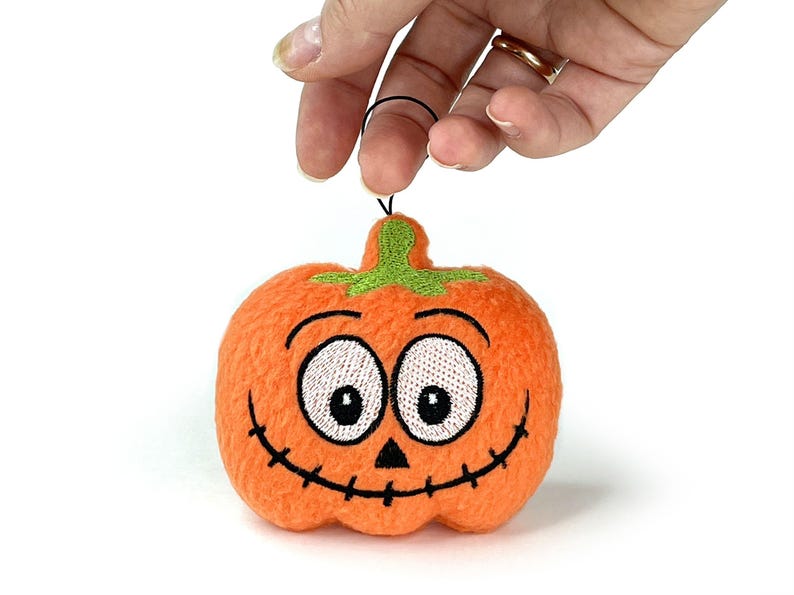 Puede incluir: Un adorno de calabaza naranja de felpa con textura difusa, que presenta ojos blancos bordados con pupilas negras, una peque&ntilde;a nariz triangular negra y una boca ancha cosida en negro. Un tallo verde bordado se encuentra en la parte superior.