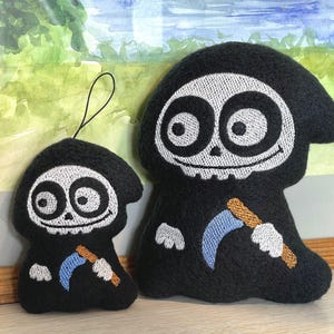 Puede incluir: Dos peluches con forma de parcas de dibujos animados, ambos con t&uacute;nicas negras con capucha, caras de calavera blancas con grandes ojos circulares y una peque&ntilde;a guada&ntilde;a azul claro con mango color canela. El peluche m&aacute;s peque&ntilde;o incluye un lazo negro.