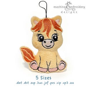 Può includere: Un cavallo di peluche beige chiaro con criniera e coda arancioni, dettagli ricamati e un viso sorridente. Il cavallo ha un cordino nero per appenderlo e il testo "machine embroidery designs" è visibile. È presente anche il testo "5 Sizes".