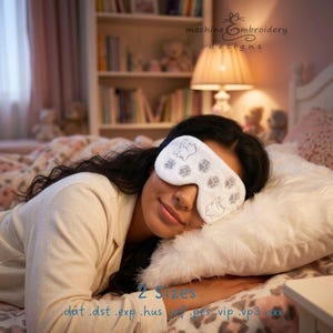 May include: A white sleep mask featuring embroidered gray cat silhouettes and paw prints. The mask is worn over the eyes, covering the upper face. The text "machine embroidery designs" appears in the upper right, with "2 Sizes" and file formats below.