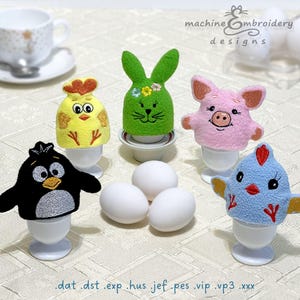 Könnte beinhalten: Fünf bestickte Eierwärmer in verschiedenen Tierdesigns: ein Küken, ein Häschen, ein Schweinchen, ein Pinguin und ein Vogel. Die Eierwärmer stehen auf weißen Eierbechern und sind von weißen Eiern umgeben. Der Text "machine embroidery designs" ist oben im Bild zu sehen.