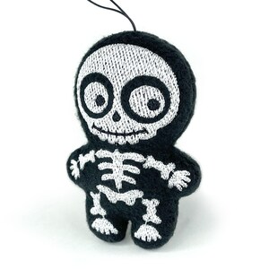 Puede incluir: Un peque&ntilde;o adorno de peluche negro con forma de esqueleto que presenta detalles bordados en blanco de un cr&aacute;neo con ojos grandes y una caja tor&aacute;cica. La figura tiene cabeza y cuerpo redondeados, con un cord&oacute;n negro fino para colgar.