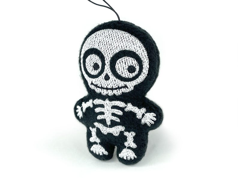 Puede incluir: Un peque&ntilde;o adorno de peluche negro con forma de esqueleto que presenta detalles bordados en blanco de un cr&aacute;neo con ojos grandes y una caja tor&aacute;cica. La figura tiene cabeza y cuerpo redondeados, con un cord&oacute;n negro fino para colgar.