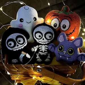 Puede incluir: Una colección de cinco adornos de peluche de Halloween en una cesta de madera, que incluye un fantasma blanco, una calabaza naranja, una parca negra, un esqueleto blanco y un murciélago morado, iluminados por una guirnalda de luces LED.