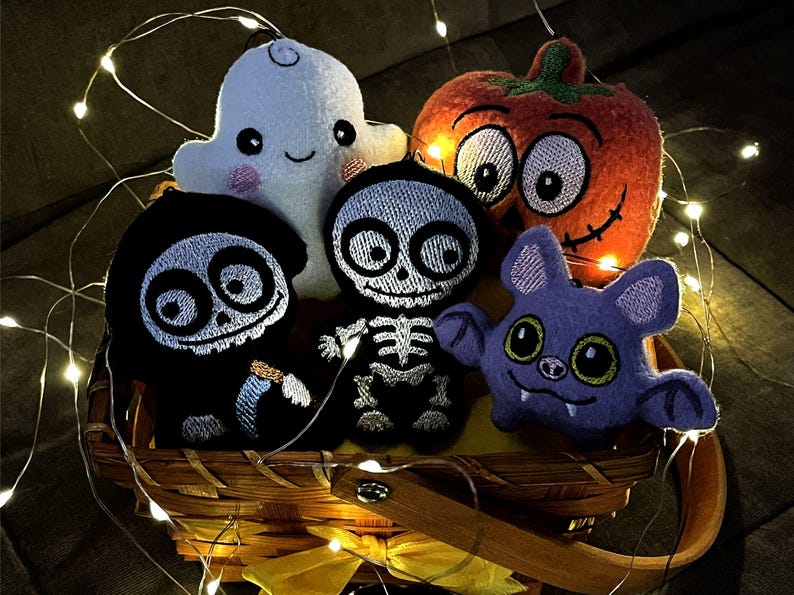 Puede incluir: Una colecci&oacute;n de cinco adornos de peluche de Halloween en una cesta de madera, que incluye un fantasma blanco, una calabaza naranja, una parca negra, un esqueleto blanco y un murci&eacute;lago morado, iluminados por una guirnalda de luces LED.