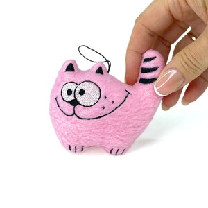 May include: A pink plush cat toy with embroidered details. The cat has a smiling face, black whiskers, and black stripes on its tail. A black string is attached for hanging.
