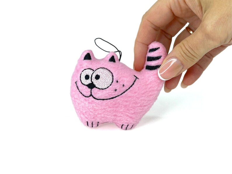 May include: A pink plush cat toy with embroidered details. The cat has a smiling face, black whiskers, and black stripes on its tail. A black string is attached for hanging.