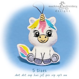 Puede incluir: Adorno de unicornio de peluche blanco con cuerno dorado, melena y cola coloridas. El unicornio tiene ojos grandes y está sentado. El texto "machine embroidery designs" está en la parte superior y "5 Sizes" en la inferior.