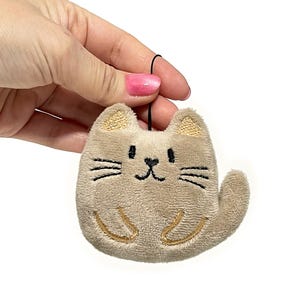 May include: A plush cat-shaped ornament in a light brown color. The cat has embroidered details, including a smiling face, whiskers, and paws. The ears are a darker shade of brown. A black string is attached for hanging.